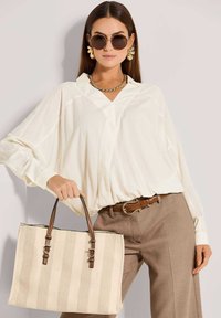 Gestreifte beige Tragetasche mit braunen Ledergriffen, getragen von einem Modell in einer cremefarbenen Bluse und braunem Gürtel, das ein lässiges, strukturiertes Design präsentiert.