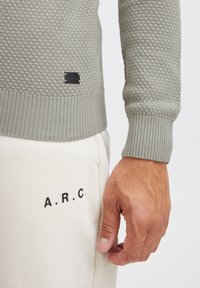 Lichtgroene, gestructureerde trui met een honingraatpatroon, geribde manchetten en een zwart label. Gecombineerd met crèmekleurige broek met zwarte letters "A.R.C."