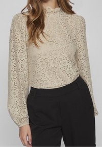 Donna che indossa una blusa lunga in pizzo color crema con colletto alto, infilata in pantaloni neri su misura.