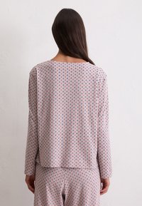 Femme faisant face à un mur blanc, portant un haut à manches longues rose et un pantalon assorti avec un petit motif géométrique sombre.