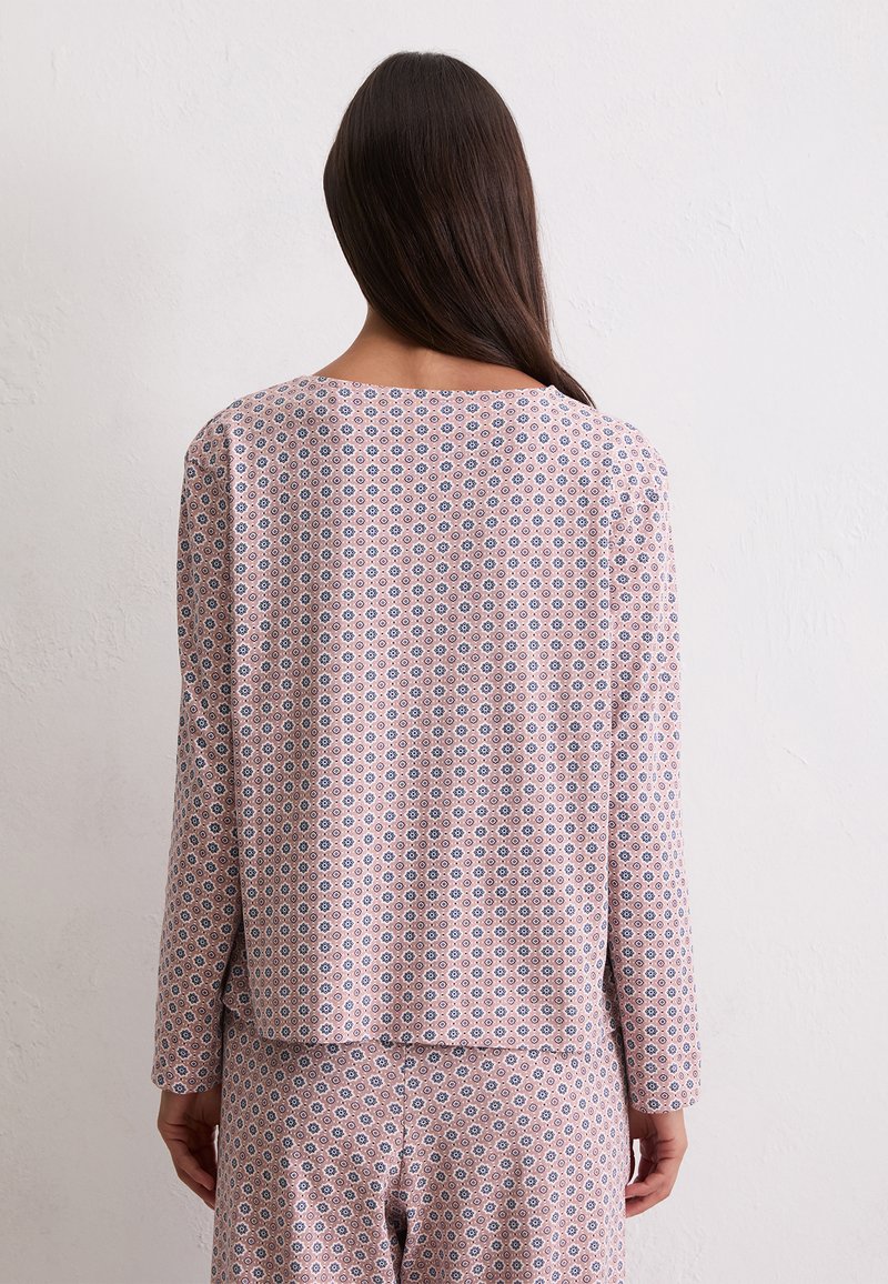 Femme faisant face à un mur blanc, portant un haut à manches longues rose et un pantalon assorti avec un petit motif géométrique sombre.