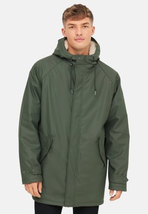 Un joven lleva una parka impermeable verde con capucha y forro de forro polar, de pie frente a un fondo blanco liso.