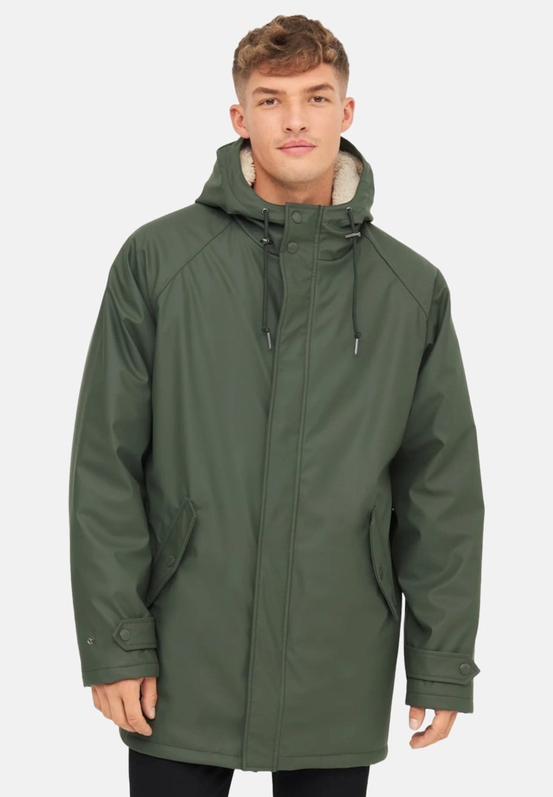 Mladý muž má na sobě zelenou nepromokavou parku s kapucí a fleecovou podšívkou, stojí na čistě bílém pozadí.