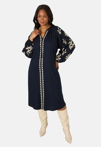 Navyblauwe jurk met geborduurde florale ontwerpen, knoopsluiting en lange pofmouwen; gecombineerd met crème knie-hoge laarzen.