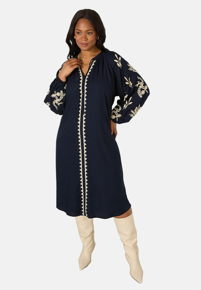 Navyblauwe jurk met geborduurde florale ontwerpen, knoopsluiting en lange pofmouwen; gecombineerd met crème knie-hoge laarzen.