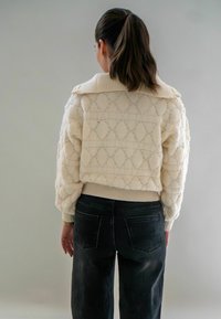 Crème gebreide trui met een gestructureerd diamanten patroon, een oversized kraag en geribbelde mouwuiteinden, gecombineerd met zwarte jeans. Zicht van achteren.