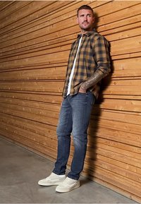 Rutig skjorta i brun och gul, lager på en vit t-shirt, kombinerad med mörka jeans och vita sneakers. Tatueringar synliga på underarmen.