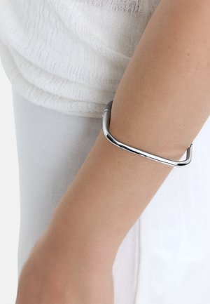 Metalowa bransoletka w formie bangla z gładkim, polerowanym wykończeniem w kolorze srebrnym, charakteryzująca się minimalistycznym, geometrycznym wzornictwem i otwartą formą.