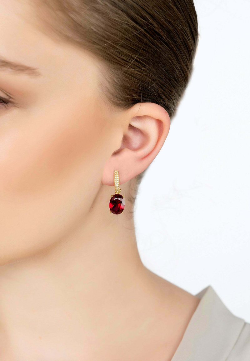 Boucles d'oreilles en or en forme de cerceau, ornées d'une grande pierre précieuse rouge ovale, avec un bandeau texturé et un fermoir sécurisé, présentées sur un fond clair.