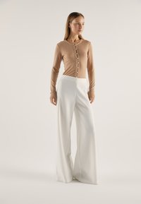 Beige ribbet cardigan med knappelukking, sammen med høytlivede, vide hvite bukser. Glatt tekstur og rene linjer gjennom hele antrekket.