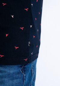 Námořnické tričko z lehkého bavlněného materiálu s malými vzory velrybích ocasů v červené a béžové barvě, boční štítek viditelný na lemu.