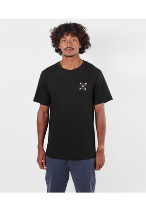 Homme aux cheveux bouclés portant un t-shirt noir avec un petit motif floral pastel symétrique sur la poitrine gauche et un pantalon bleu.