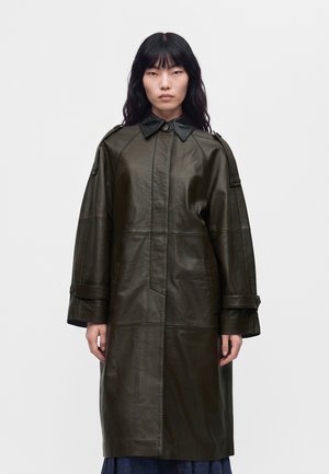 Le modèle se tient face à l’objectif, vêtu d’un long manteau en cuir vert olive foncé, avec un col noir et des manches larges, sur un fond clair et uni.
