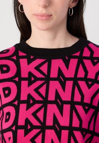 Fuksijos megztinis su ryškiais juodais "DKNY" užrašais, geometriniais raštais ir juodu nertiniu apykakle. Modelis vilki sidabrinius žiedus ausyse.
