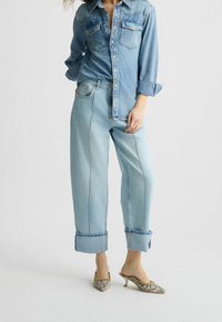Camisa de mezclilla azul claro con botones en el frente y dos bolsillos en el pecho, combinada con pantalones vaqueros de pierna ancha azul claro, con puños enrollados y mules de tacón con estampado.