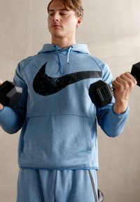 Kék kapucnis pulóver nagy fekete Nike logóval, passzoló kék nadrággal párosítva, fekete kézisúlyzókat emel egy semleges háttérben.