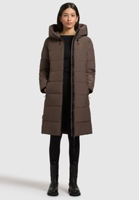 Langer brauner Daunenmantel mit Kapuze, mit gesteppten Abschnitten, einem Reißverschluss an der Vorderseite und Taschen. Getragen über schwarzen Leggings und Ankle Boots.