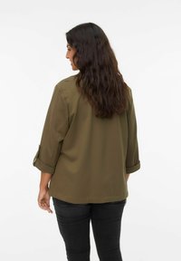 Olivgrüne Bluse mit lockerer Passform, Dreiviertel-Ärmeln und glatter Textur. Hat einen geraden Saum. Schwarze Jeans sind sichtbar.