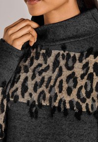 Nahaufnahme einer Person, die am Kragen eines dunkelgrauen Pullovers zieht, der ein beige und schwarzes Leopardenmuster auf der Brust hat.