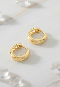 Guldörrhängesmycken med en slät yta och små klara stenar som accent. Uppställda på en marmorlika bakgrund med spridda klara stenar.