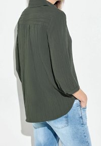 Chemise boutonnée vert olive, texturée, avec un col et des manches trois-quarts, portée avec un jean bleu clair.