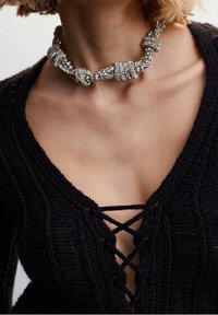 Srebrna choker z koralikami o węzełkowym wzorze. Noszona na czarnej dzianinowej bluzce z głębokim dekoltem w kształcie V i krzyżującymi się detalami.