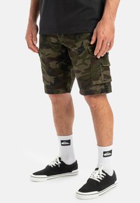 Pantalones cortos cargo de camuflaje en tonos verdes y marrones, con bolsillos laterales, un ajuste relajado y una construcción de tela duradera. Calzado negro y calcetines blancos.