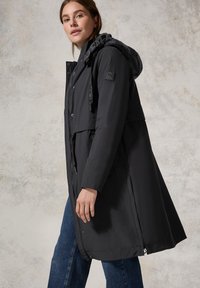 Parka nera impermeabile con cappuccio texturizzato, vita cinchata e tasche laterali. Presenta una chiusura con zip e un dettaglio con logo sulla manica.