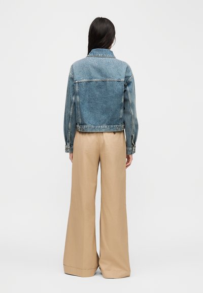 Γυναίκα με μακριά σκουρόχρωμα μαλλιά, φορώντας ένα μπλε τζην μπουφάν και μπεζ παντελόνι με φαρδιά πόδια, που στέκεται γυρισμένη πλάτη μπροστά σε ένα απλό φόντο.