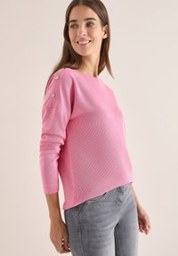 Rosa Strickpullover mit drei Viertelärmeln, der Metallknopf-Akzente an den Schultern und einen lässigen Schnitt aufweist. Kombiniert mit hellgrauen Jeans.