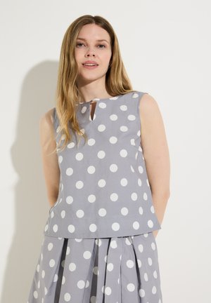 Woman with long hair wearing sleeveless gray top and skirt with large white polka dots, standing against a plain light background.