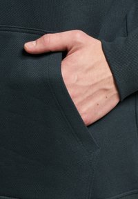 Dunkelgrüne strukturierte Kapuzenjacke mit einer Kängurutasche, die einen engen Schnitt und gerippte Bündchen aufweist. Eine Hand befindet sich in der Tasche.