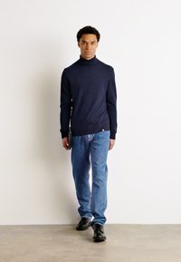 Ναυτικό ζιβάγκο, πλεκτό ύφασμα, εφαρμοστή σχεδίαση; συνδυασμένο με ανοιχτό μπλε τζιν και μαύρα παπούτσια, κρατώντας ένα μαύρο αντικείμενο.