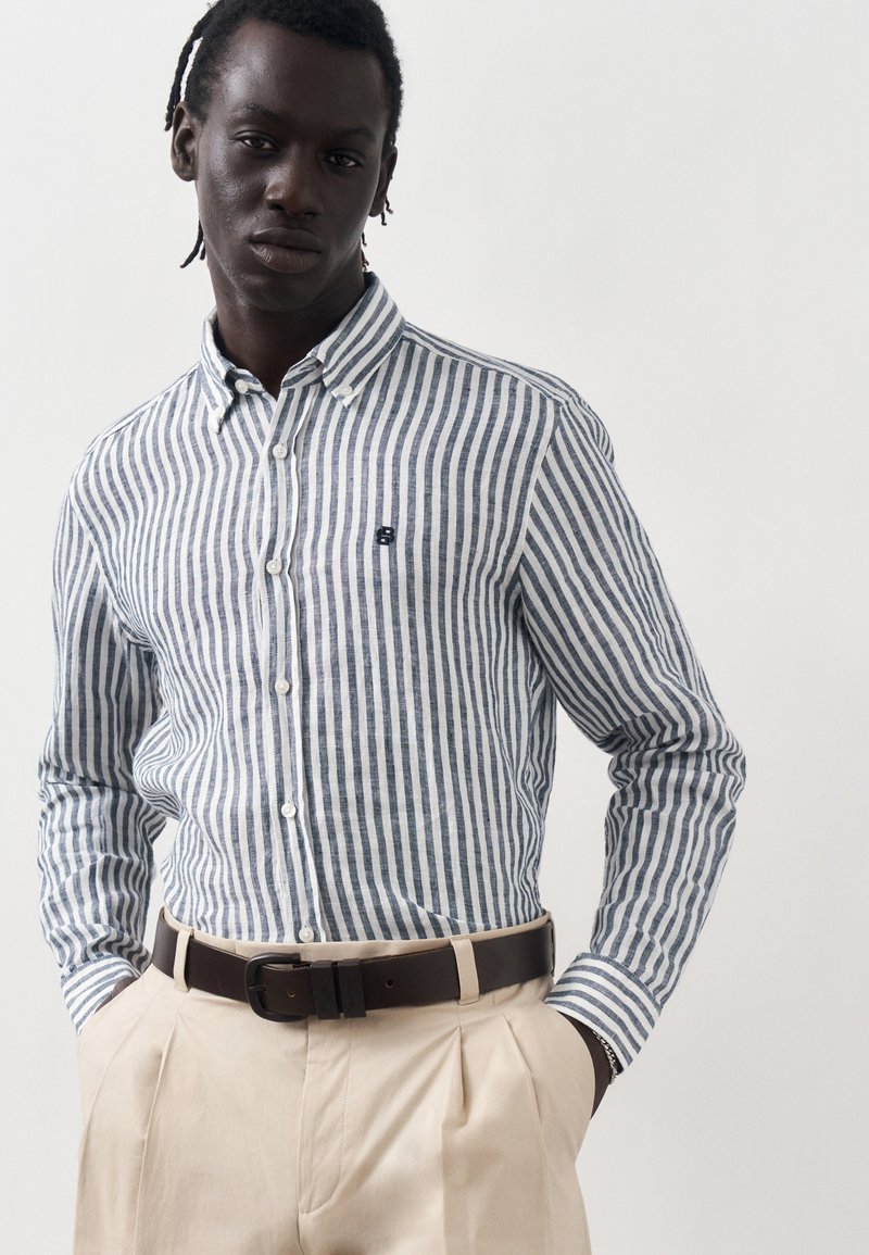 Άνδρας φοράει μπλε και λευκό πουκάμισο με κάθετες ρίγες, μπροστά κουμπωμένο και μαζεμένο μέσα σε μπεζ πλισέ παντελόνια με σκούρα καφέ ζώνη, τα χέρια στις τσέπες.