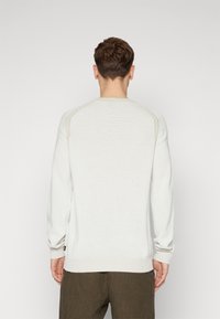 Jasnokremowy sweter typu pulower z prążkowanymi mankietami i dolnym wykończeniem, z fakturowanym wzorem dzianiny i kontrastowymi szwami na ramionach. Noszony z kratkowymi spodniami.