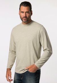 Camiseta de manga larga beige claro, hecha de tela suave, con cuello redondo, que presenta un sutil logo en el pecho, combinada con jeans oscuros.