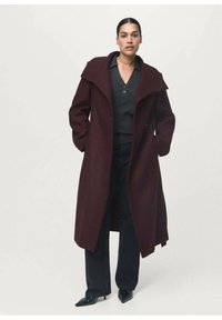 Long manteau bordeaux avec un large col, porté sur un haut noir et un pantalon foncé. Le tissu est lisse avec une coupe ajustée et des poches latérales.