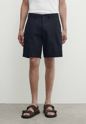 Homme portant un short noir jusqu'aux genoux, un t-shirt blanc et des sandales marron foncé, debout sur un sol gris contre un mur blanc.