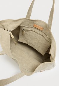 Sac fourre-tout en tissu beige avec deux poches zippées, une doublure intérieure et une étiquette en cuir. Design simple et spacieux avec des poignées solides.