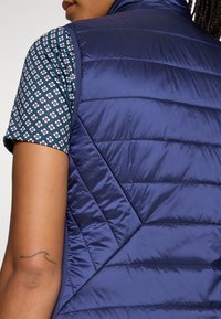 Marineblaues Puffer-Weste mit gestepptem Design und glatter Textur. Darunter ein gemustertes kurzärmeliges Hemd in dunklen Farben mit floralen Motiven.