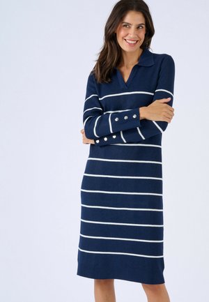 Mujer sonriendo con los brazos cruzados, vistiendo un vestido suéter azul marino con rayas horizontales blancas y botones plateados en las mangas.