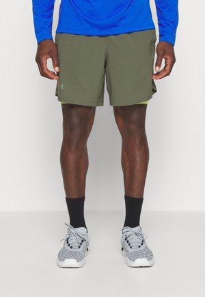 Mand stående iført olivengrønne sportsshorts, sorte sokker, grå løbesko og en blå langærmet top, som er skåret af ved midt låret.