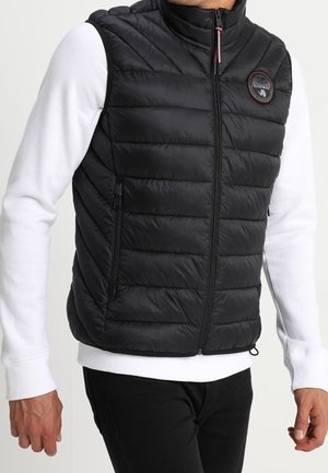 Mężczyzna w czarnej pikowanej kamizelce na zamek, na białej koszuli z długim rękawem i czarnych spodniach, stoi z rozluźnionymi rękami wzdłuż ciała.