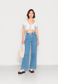 Top cropped azul claro com mangas bufantes e detalhe laço, combinado com calças de ganga largas de cintura alta azuis, sandálias de plataforma pretas e uma mala bege.