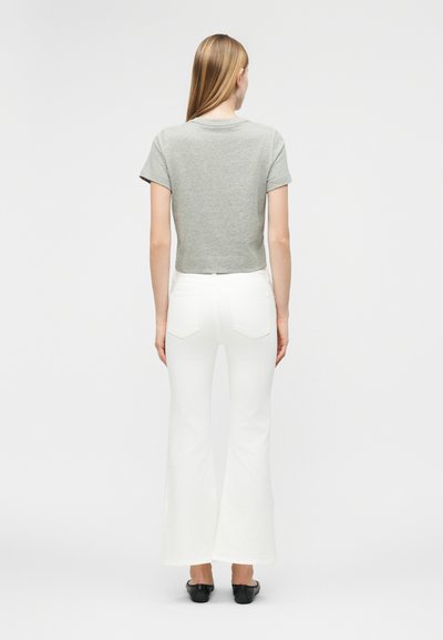 Γυναίκα με μακριά ξανθά μαλλιά που φοράει γκρι μπλουζάκι με κοντά μανίκια, λευκό καμπάνα παντελόνι και μαύρα παπούτσια, στέκεται με την πλάτη γυρισμένη σε απλό φόντο.