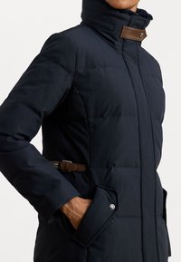 Tamsiai mėlynas pūkinis paltas su aukštu apykakle, rudi odiniai akcentai ir priekinės kišenės. Vijos tekstūra ir šoninės dirželio detalės reguliavimui.