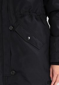 Sektion av svart vinterjacka med knappad klaffficka, stora knappar, åtsittande midja och en hand som vilar mot sidan.