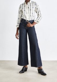 Frau trägt ein gemustertes weißes Hemd mit Knopfleiste, dunkle weit geschnittene Jeans und schwarze High-Heel-Mokassins, steht auf einem schlichten Boden.
