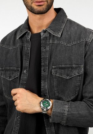 Orologio d'argento con quadrante verde, cinturino in acciaio inossidabile, indossato con una camicia di jeans grigio scuro con bottoni a pressione e tasche sul petto.
