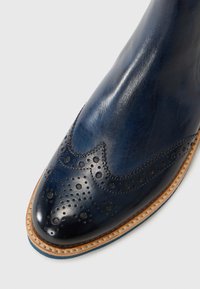 Botte cheville en cuir navy avec des détails brogue et des perforations. Présente un bout arrondi et une semelle en cuir beige cousue. Texture lisse et finition brillante.